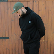 Man wearing a black Destroy or Die hoodie and green cap against a wooden wall