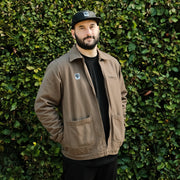 Man wearing a brown jacket and black cap standing against a green hedge