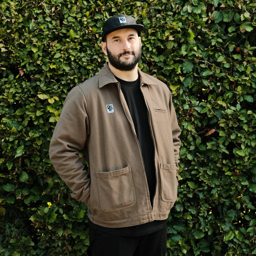 Man wearing a brown jacket and black cap standing against a green hedge