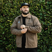Man wearing a brown jacket and black cap standing against a green hedge.