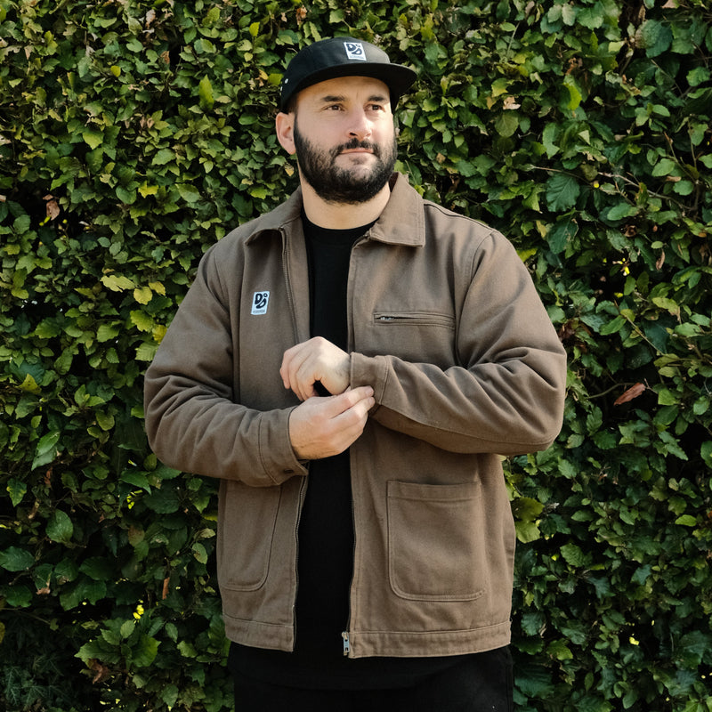 Man wearing a brown jacket and black cap standing against a green hedge.