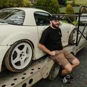 Man sitting on a tow truck with a white car behind him, outdoors.
