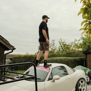 Man standing on a white car in brown shorts, outdoors on a cloudy day.