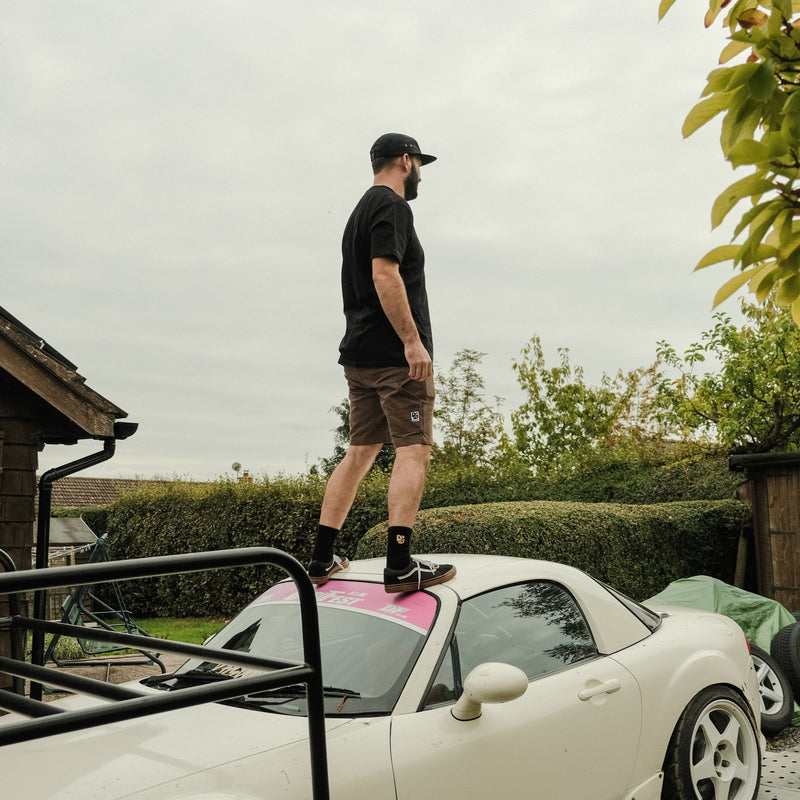 Man standing on a white car in brown shorts, outdoors on a cloudy day.