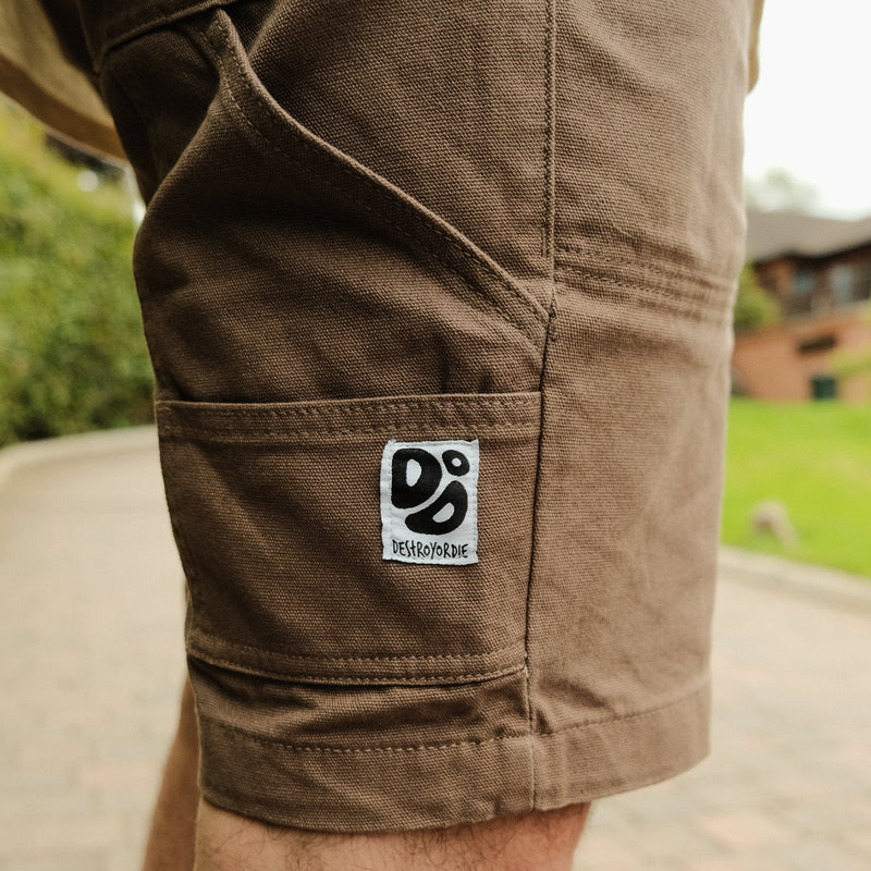Brown shorts with a  Destroy or Die logo on the pocket against a blurred outdoor background