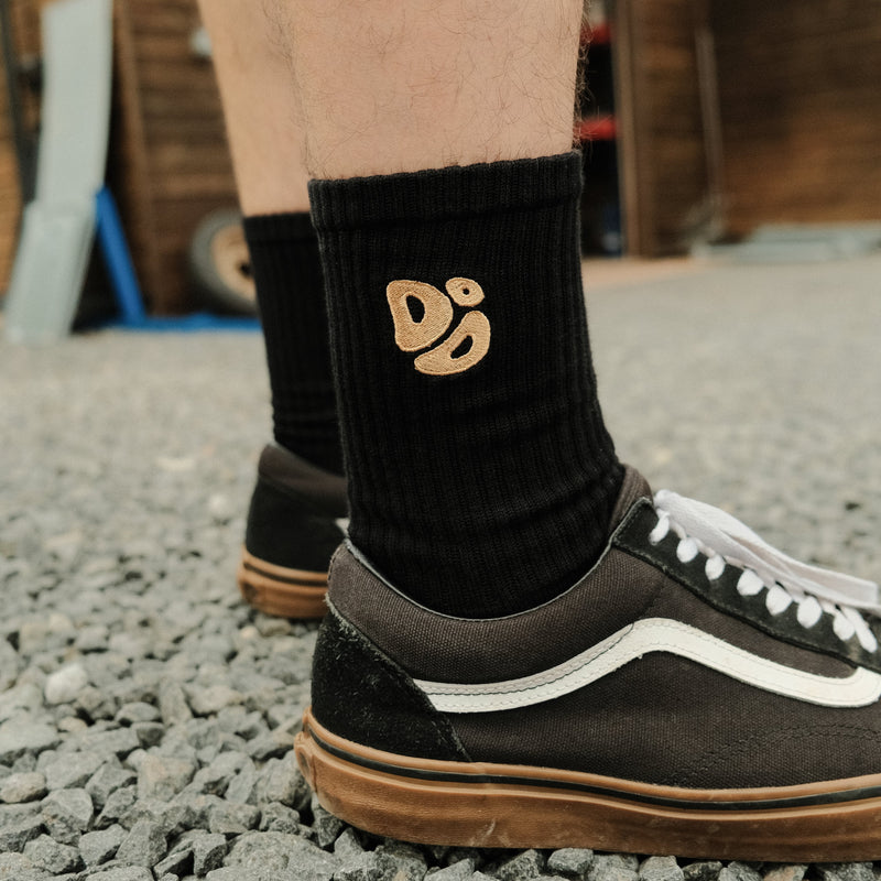 Person wearing black socks with a Destroy or Die logo and sneakers on a gravel surface.