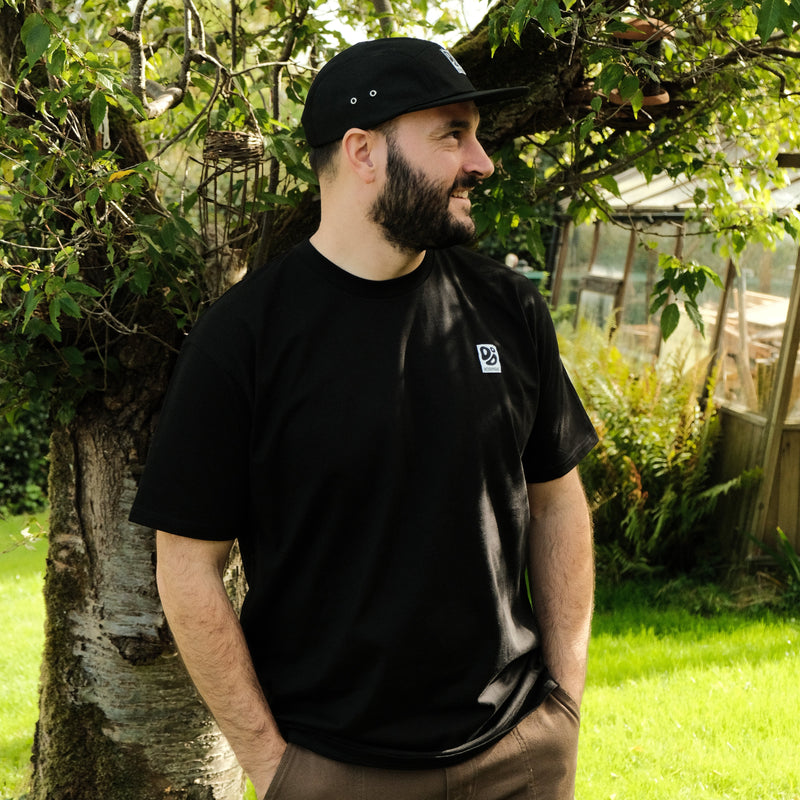 Man wearing a black t-shirt with a Destroy or Die logo standing outdoors near trees and greenery