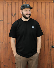 Man wearing a black t-shirt with a Destroy or Die logo and a cap, standing against a wooden fence.