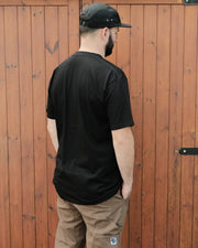 Man wearing a black t-shirt and cap standing against a wooden fence