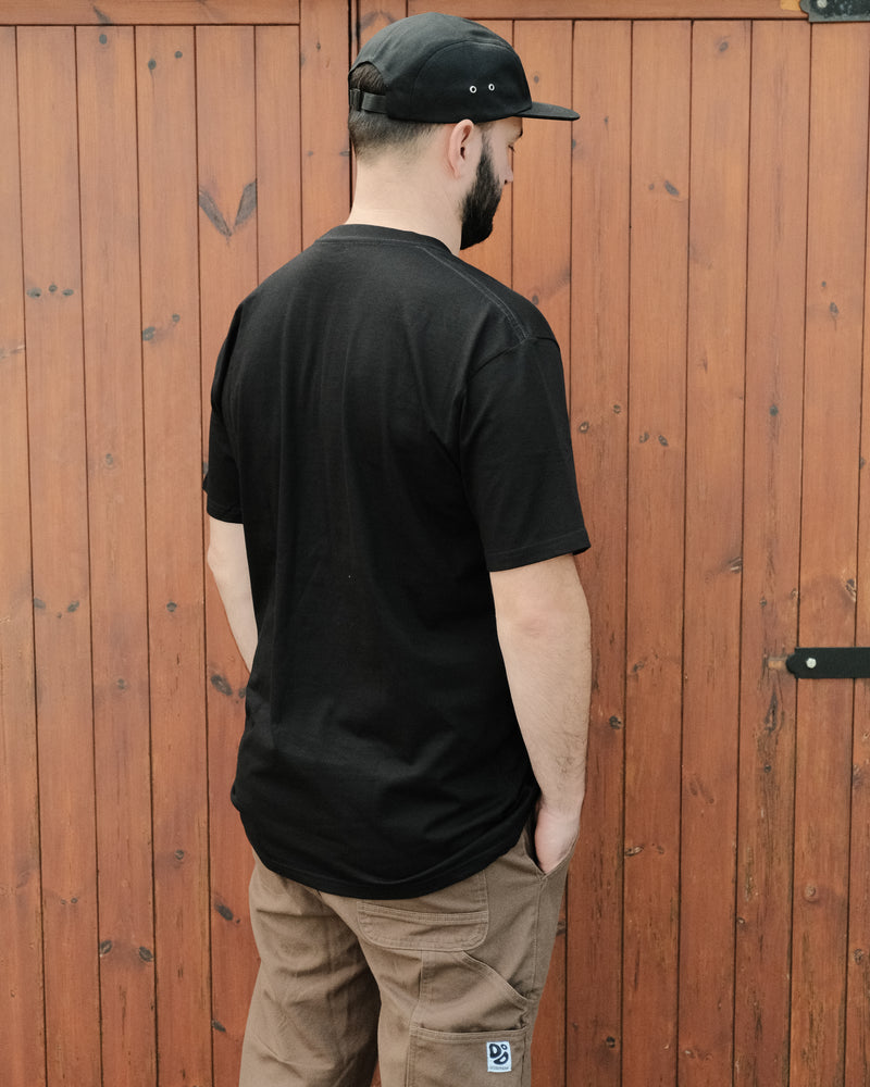 Man wearing a black t-shirt and cap standing against a wooden fence