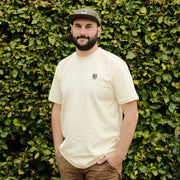 Man wearing an ecru t-shirt and cap standing against a green leafy background