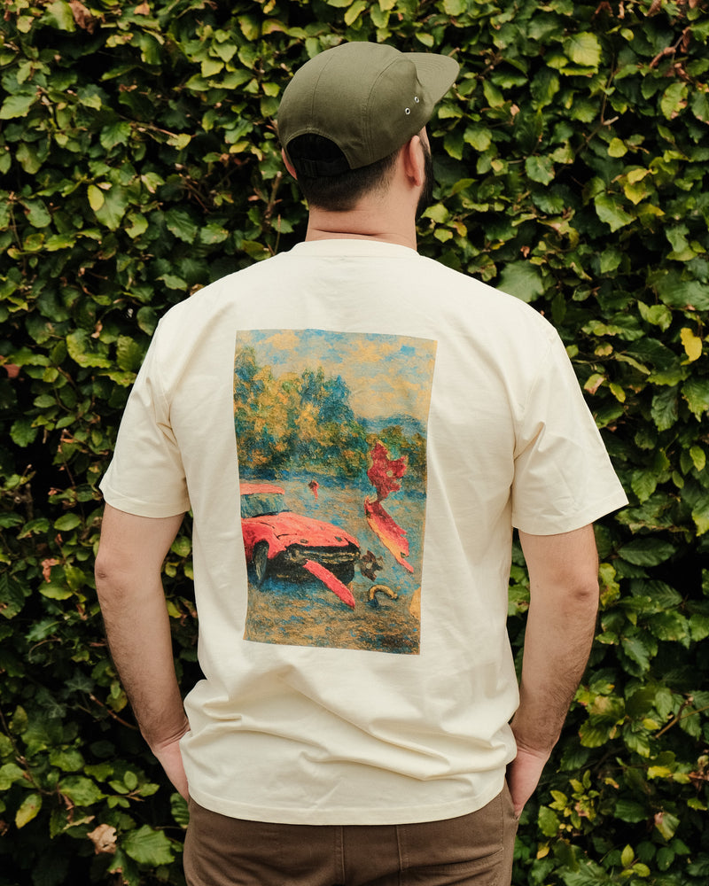Person wearing a ecru t-shirt with a graphic design on the back, standing against a green hedge.