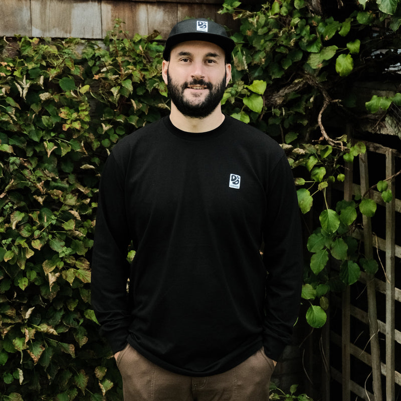 Man wearing a Destroy or Die black long-sleeve shirt and cap standing in front of green foliage.
