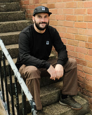 Man sitting on a set of stairs against a brick wall wearing a Destroy or Die black long sleeve t-shirt and cap.