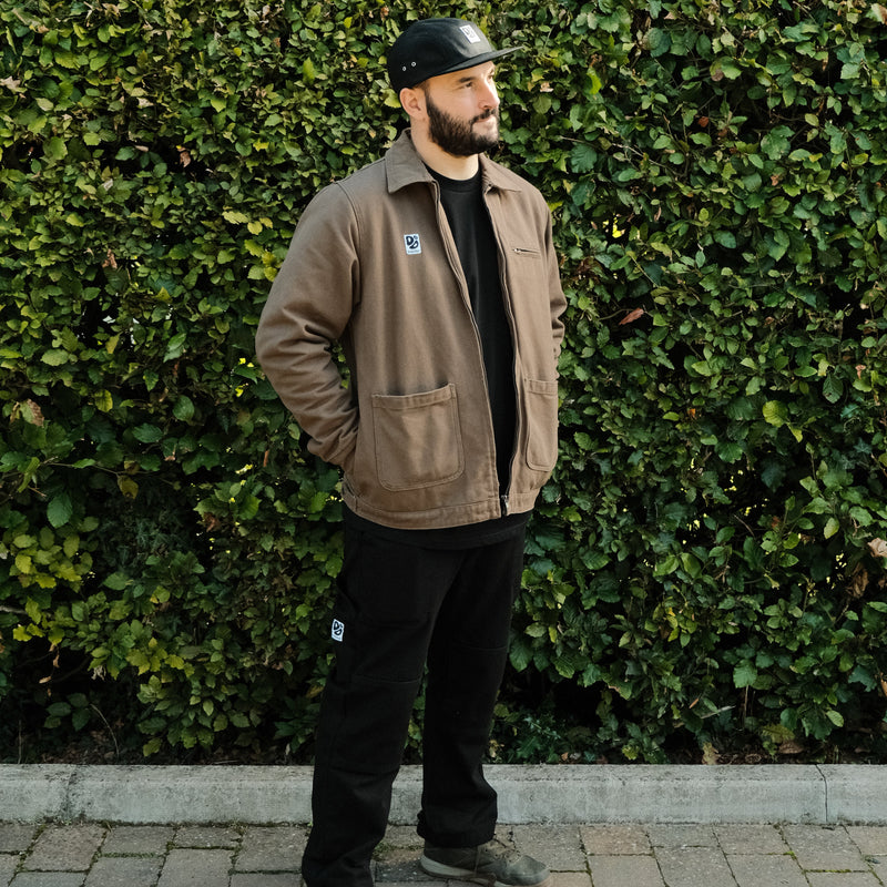 Man wearing a brown jacket and black pants standing against a green hedge.