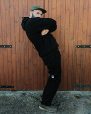 Person wearing a black hoodie and pants with a cap, standing against a wooden wall.